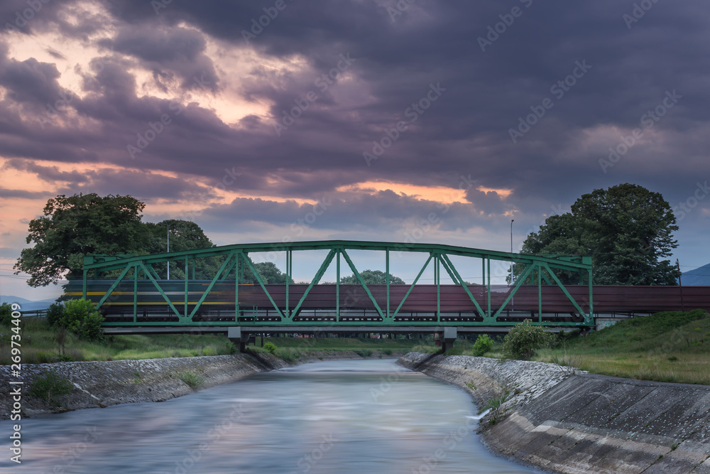 Fototapeta premium Blurred motion, long exposure of train passing over metallic railway bridge during blue hour with sky still dramatic after sunset