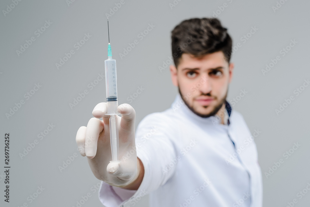 Middle eastern medical doctor holding a full suringe in focus in gloves isolated over grey background Middle eastern medical doctor holding a full suringe in focus in gloves isolated over grey background