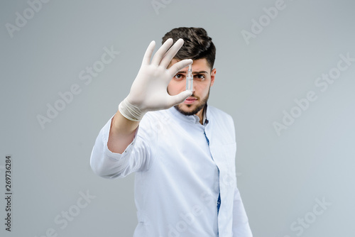 Attractive medical doctor in gloves holding an ampoule in front of him isolated over grey background photo