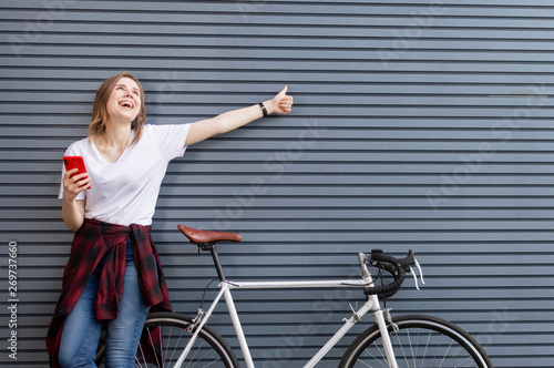 young beautiful girl stands with a bicycle on the background of a wall and looks into the phone, a woman shows a finger to a place for text