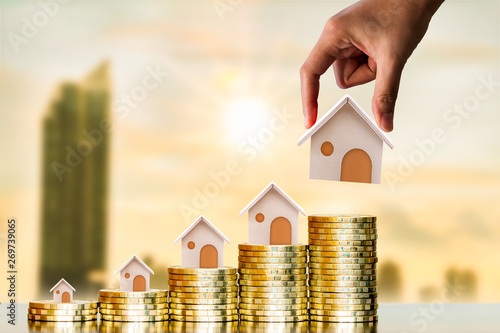 Investor hand hold a home model put on the stack coin with growing on photo blur cityscape on sunlight background, Savings money for buy house and loan to business investment for real estate concept. photo