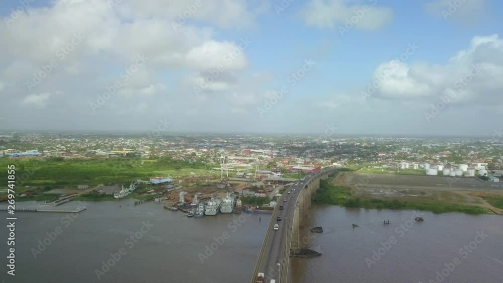 Wide angle shot of high flying drone passing over Jules Wijdenbosch Bridge between Paramaribo and Meerzorg in Suriname with panoramic view of river, port, landscape and town in background. Wide angle shot of high flying drone passing over Jules Wijdenbosch Bridge between Paramaribo and Meerzorg in Suriname with panoramic view of river, port, landscape and town in background.