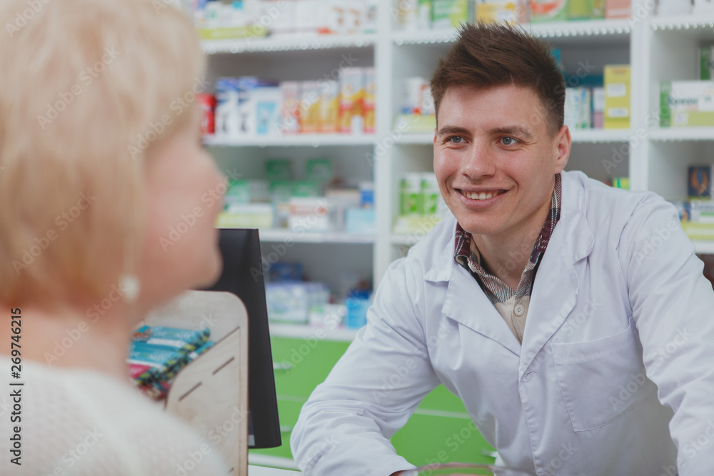 Cheerful young male chemist smiling joyfully, helping senior female customer. Rear view shot of an elderly woman shopping for medicament at local pharmacy. Retail, sales concept