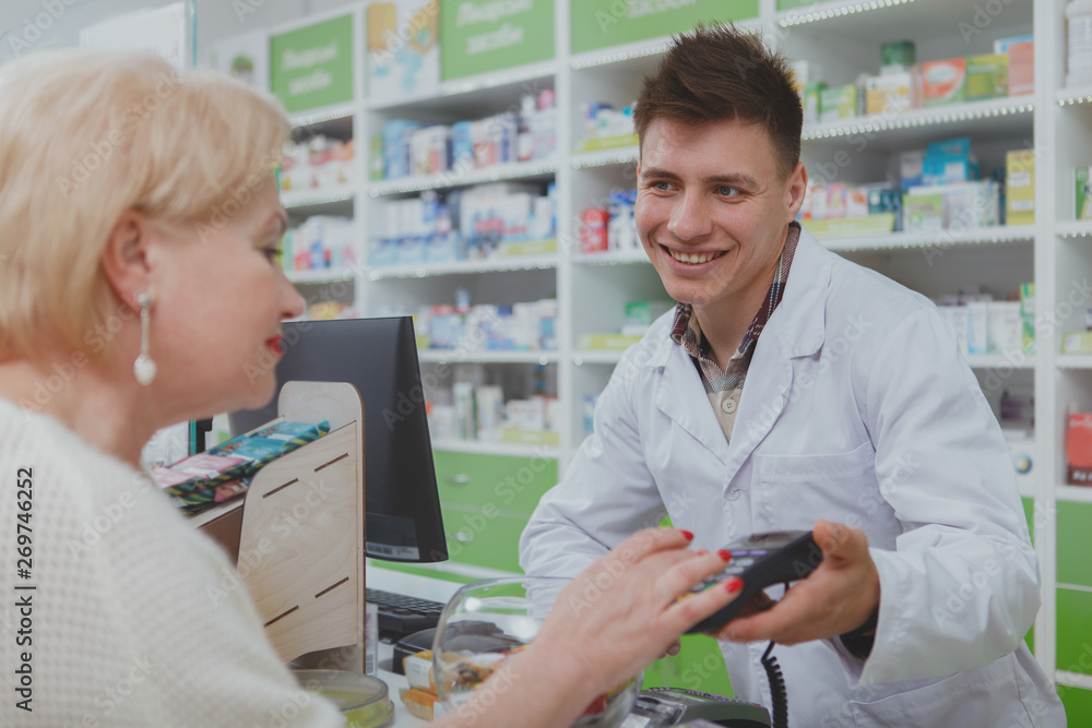 Obraz premium Senior womn paying for her purchase with a credit card at the drugstore. Cheerful male pharmacist holding card reader terminal, his elderly female customer entering security pin to pay for medicine