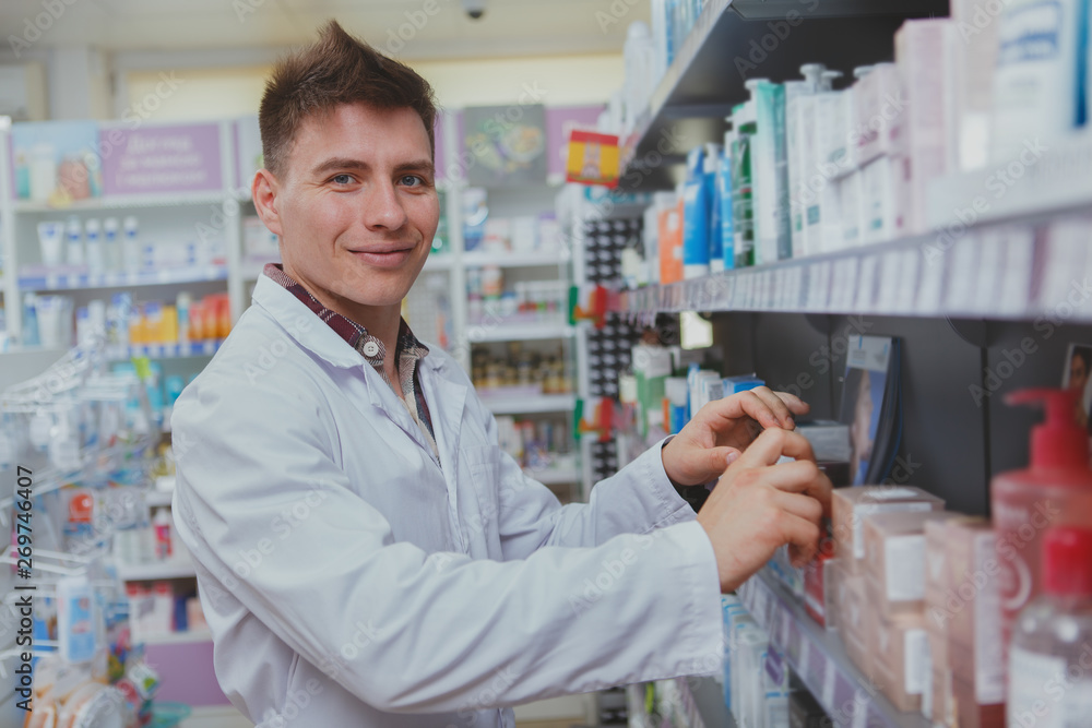 Charming male pharmacist smiling to the camera, filling shelves of drugstore with new products for sale, copy space. Cheerful chemist enjoying working at his drugstore