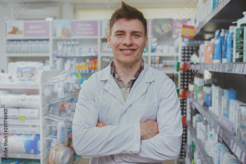 Happy handsome pharmacist standing with arms crossed at the drugstore. Friendly male chemist welcoming you in his pharmacy. Profession, occupation, medical service concept © Ihor