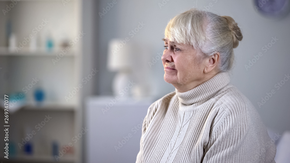 Fototapeta premium Depressed senior woman sitting alone in living room, abandoned by relatives