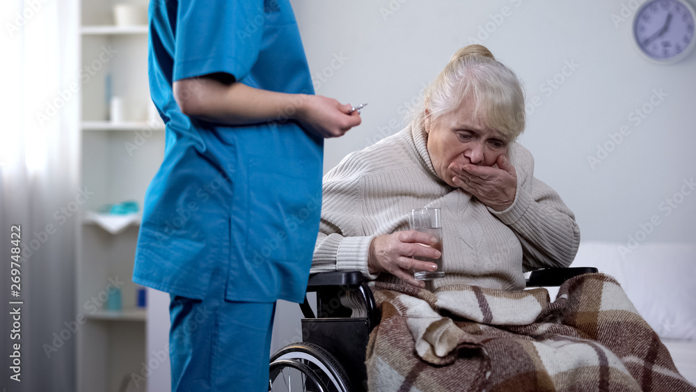Fototapeta premium Young nurse giving medications to old lady in wheelchair, rehabilitation center