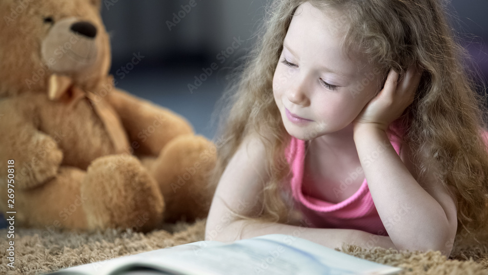 Concentrated cute girl reading book, lying on carpet, preschool education Concentrated cute girl reading book, lying on carpet, preschool education