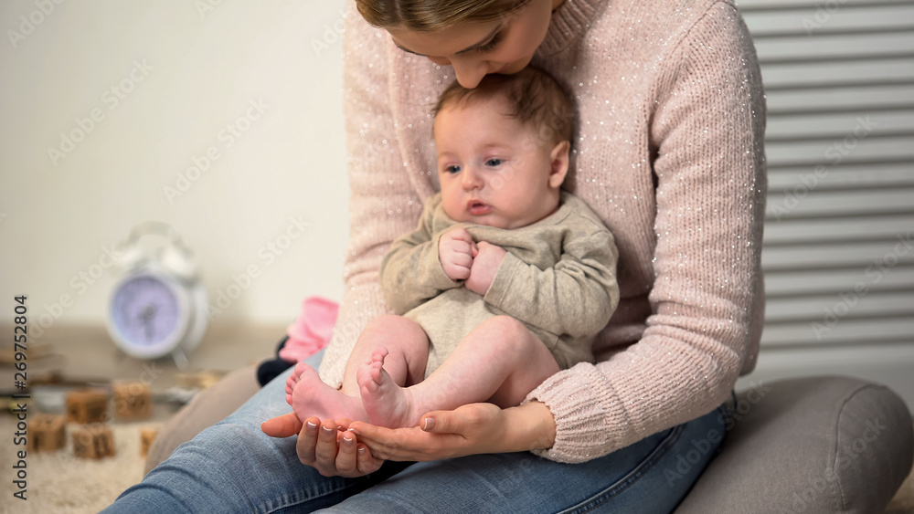Young mother admiring sons tiny legs, baby sitting on womans lap, happiness Young mother admiring sons tiny legs, baby sitting on womans lap, happiness