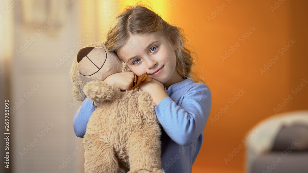 Cute curly-haired blond girl hugging teddy bear and smiling at camera, happiness - obrazy, fototapety, plakaty 