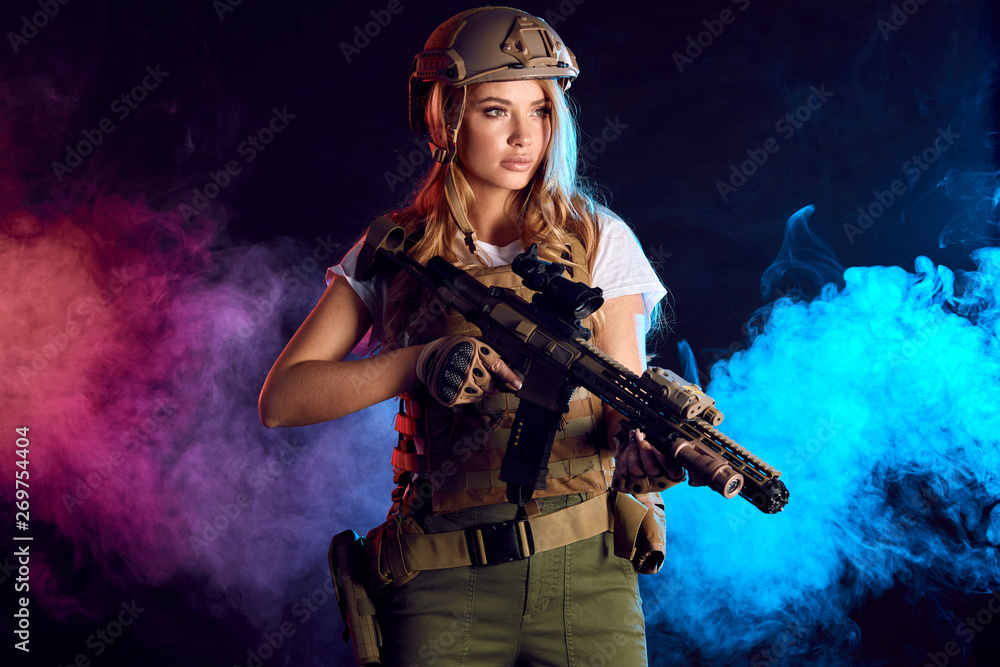 Skilled blonde female soldier with rifle in hands standing in military outfit in smoky darkness. Woman, Military Service and firearm concept