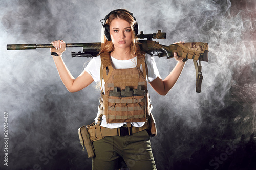 Woman soldier in military camouflage uniform protected with helmet, body armour, holding assault rifle standing against dark smoky background photo