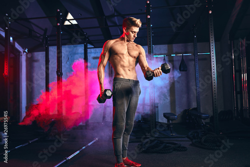 Caucasian man working out in gym doing exercises with dumbbells at triceps, strong male naked torso, highly defined muscles, six-packs slim trained body in dark gym with red spotlights photo