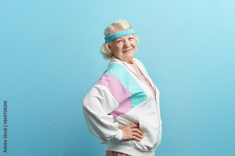 European retired woman in sportive clothings and headband, having beaming with joy face expression stands isolated in studio over blue background, copy space European retired woman in sportive clothings and headband, having beaming with joy face expression stands isolated in studio over blue background, copy space