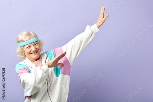 European retired woman in sportive clothings and headband, having beaming with joy face expression gesturing with hands while posing isolated in studio over blue background, copy space photo