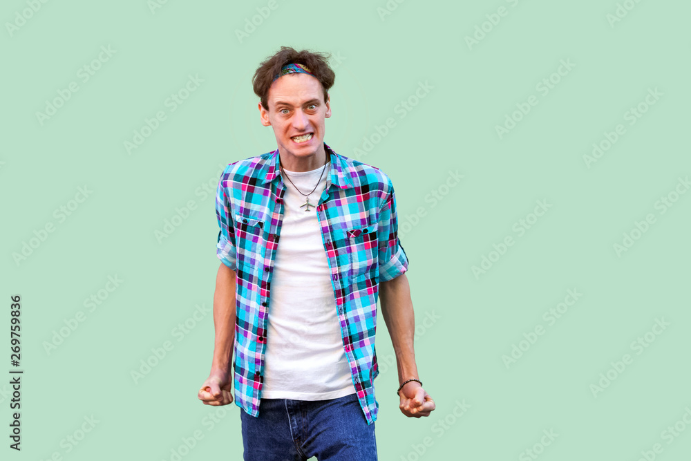 Portrait of angry nervous young man in casual blue checkered shirt headband standing, clenching teeth, looking at camera with mad furious face. indoor studio shot, isolated on light green background. Portrait of angry nervous young man in casual blue checkered shirt headband standing, clenching teeth, looking at camera with mad furious face. indoor studio shot, isolated on light green background.