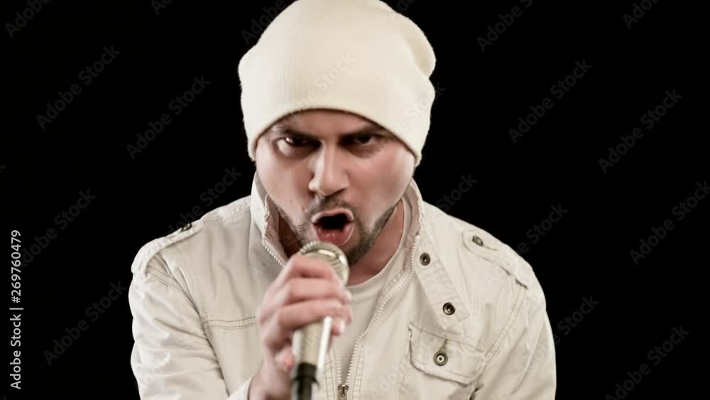 Close-up Frontman vocalist rock pop with a stylish beard in white clothes and a hat with a microphone in his hands expressively aggressively singing in the studio against the background of black walls Close-up Frontman vocalist rock pop with a stylish beard in white clothes and a hat with a microphone in his hands expressively aggressively singing in the studio against the background of black walls