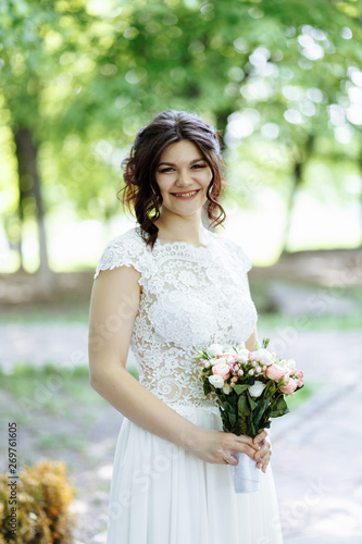 .Beautiful happy fiancee in long white dress with bouquet in hands look at camera and smiling in summer park.