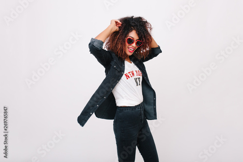 Slim girl with curly hairstyle laughing while dancing in studio. Charming tanned young lady in sunglasses and denim jacket having fun on white background.