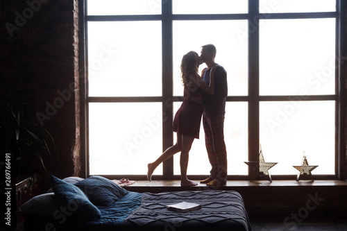 Silhouette of young happy beautiful couple expecting baby standing together in the big window sill hugging and kissing each other with love. Studio shot  love story  indoor