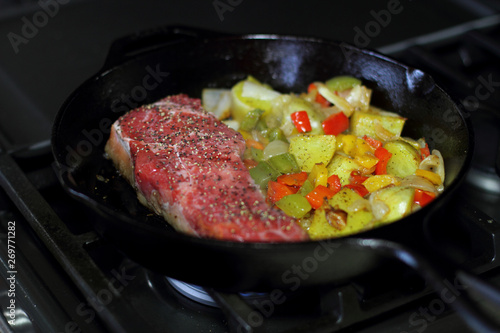 New York strip steak frying in a cast iron pan with potatoes,assorted bell peppers, and onions on the stove top.