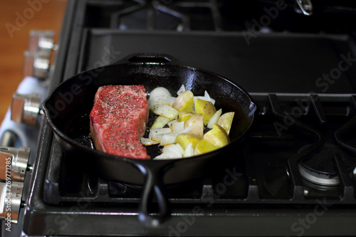 New York strip steak frying in a cast iron pan with potatoes and onions on the stove top.