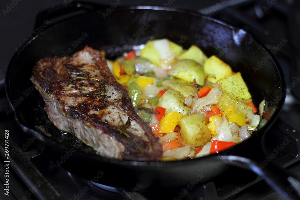 New York strip steak frying in a cast iron pan with potatoes,assorted bell peppers, and onions on the stove top.