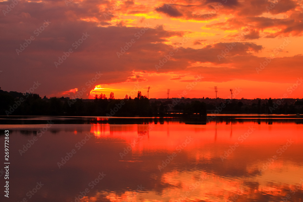 Beautiful dark sunset on the river with symmetry of colorful clouds cumulonimbus. Golden orange reflection of the sun's rays in water. Isolated tower on the skyline