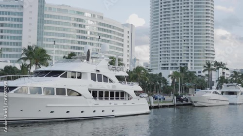 Luxury yachts parked in a Miami dock with a road with cars, palm trees and skyscrapers in the background under a cloudy sky in summer. 4K. Taken from a boat.