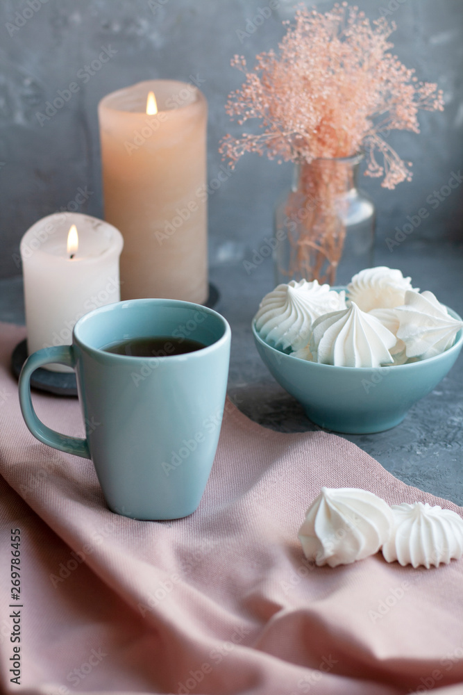 A hot cup of black tea with a bowl of airy meringues on a pink tablecloth, wax candles, a glass vase with decorative herbs on a gray background.