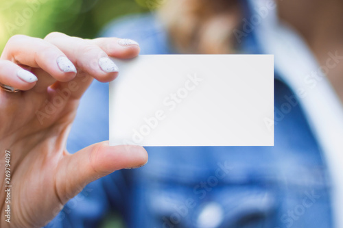 The girl holds in her hand a business card a mock up for the presentation of the design. Female hand holding a white business card with copy space on a green background of nature