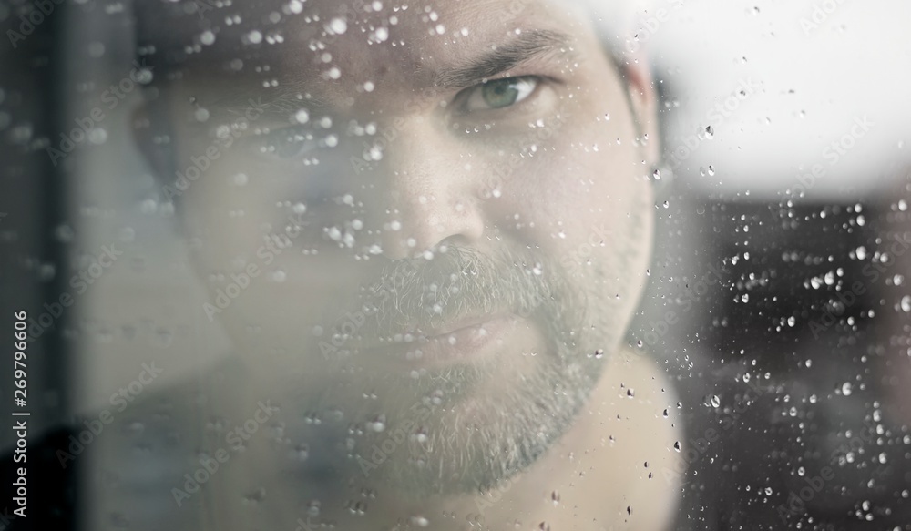 Portrait of a man looking sad through a wet window to the camera.