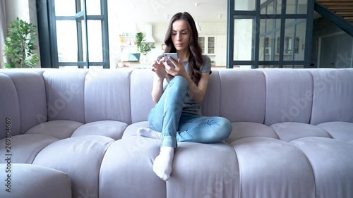 Excited and emotional young beautiful brunette woman with smart phone in hands is shouting and joy while sitting on the couch at home. Very happiness girl.