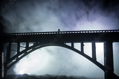 Fototapeta Naklejka Na Ścianę i Meble -  Artwork decoration. Silhouette of powerful metallic bridge at night with foggy backlight. Silhouette of person standing on bridge.