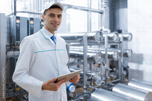 Portrait of man in a white robe and a cap standing in production department of dairy factory with grey tablet