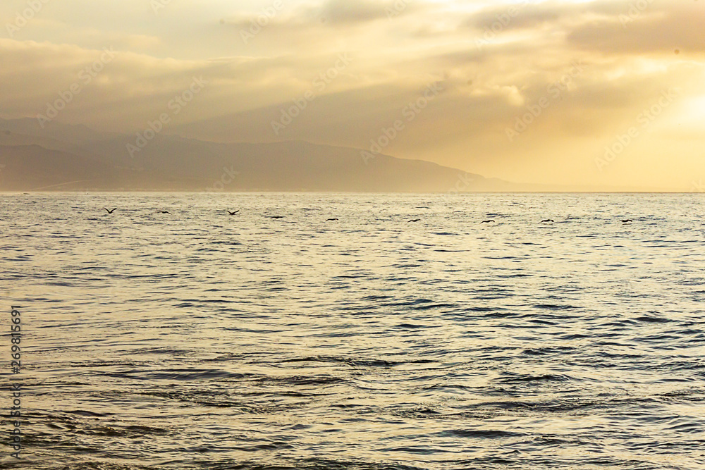 Obraz premium open ocean expanse at sunrise, with clouds, sunrays, water texture, and distant hills,