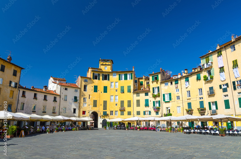 Piazza dell Anfiteatro square in circus yard of medieval town Lucca historical centre, old colorful buildings with shutter windows and blue clear sky copy space background, Tuscany, Italy