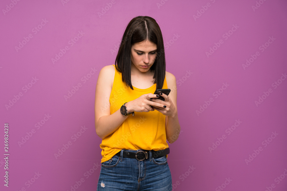 Young woman over isolated purple wall sending a message with the mobile Young woman over isolated purple wall sending a message with the mobile