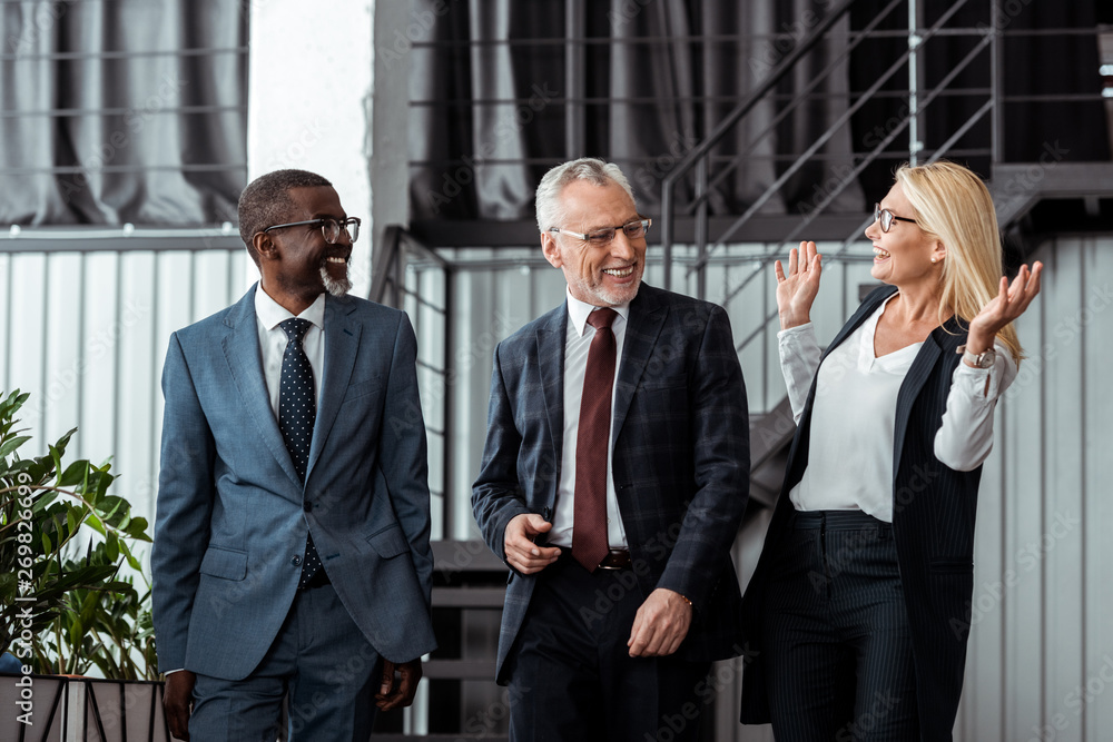 happy businesswoman gesturing while smiling with multicultural partners in office happy businesswoman gesturing while smiling with multicultural partners in office