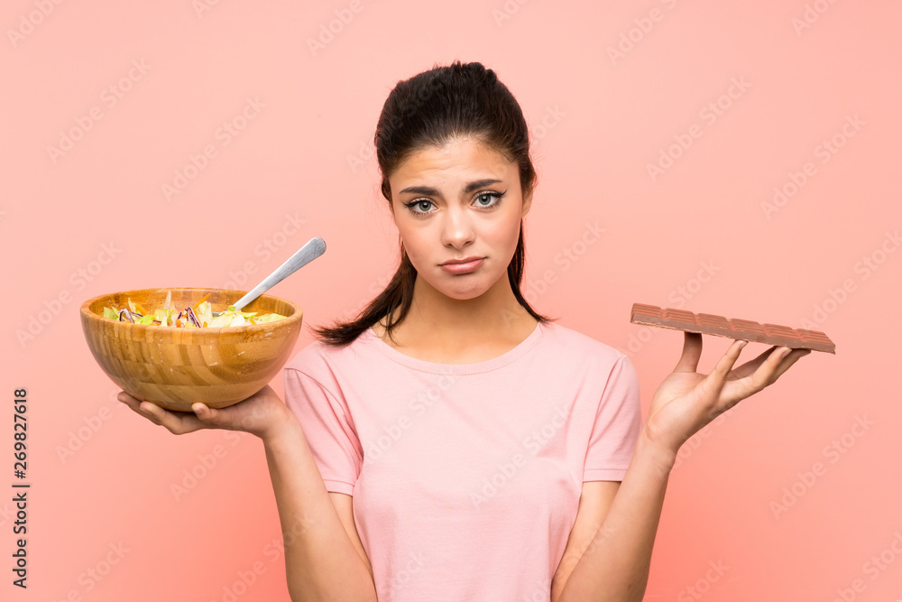 Teenager girl over isolated pink wall with salad and chocolat and having doubts Teenager girl over isolated pink wall with salad and chocolat and having doubts