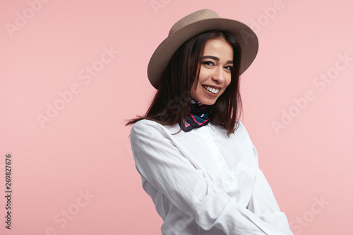 Young beautiful brunette girl wearing elegant hat, white shirt and stylish scarf, smiling and standing against pink wall