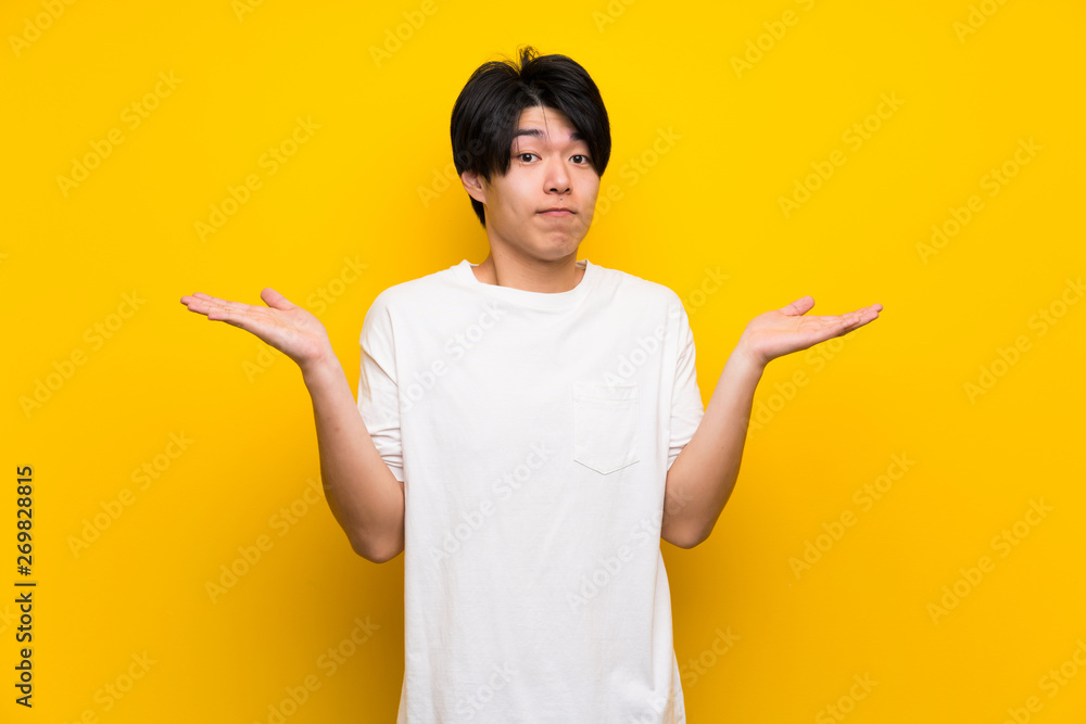 Asian man over isolated yellow wall making doubts gesture Asian man over isolated yellow wall making doubts gesture