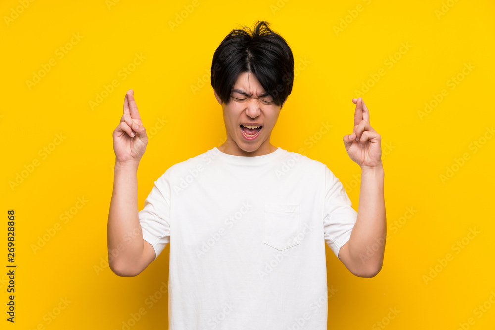 Asian man over isolated yellow wall with fingers crossing