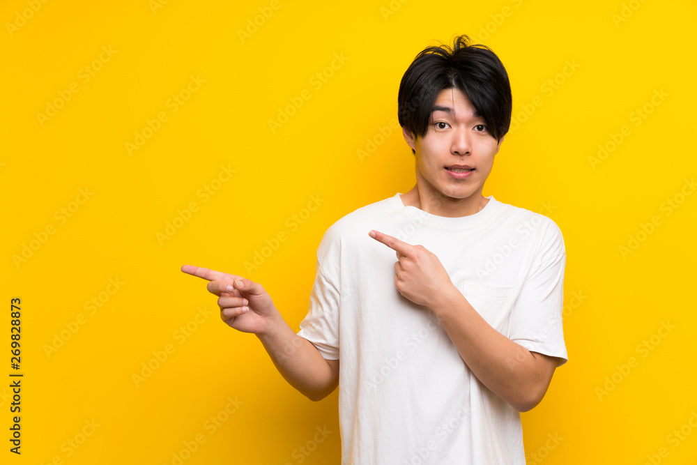 Asian man over isolated yellow wall frightened and pointing to the side Asian man over isolated yellow wall frightened and pointing to the side