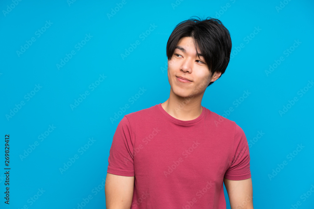 Asian man with red shirt over isolated blue wall standing and looking to the side