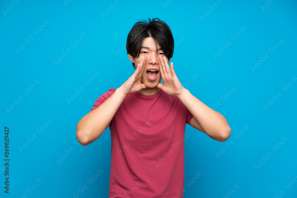 Asian man with red shirt over isolated blue wall shouting with mouth wide open Asian man with red shirt over isolated blue wall shouting with mouth wide open