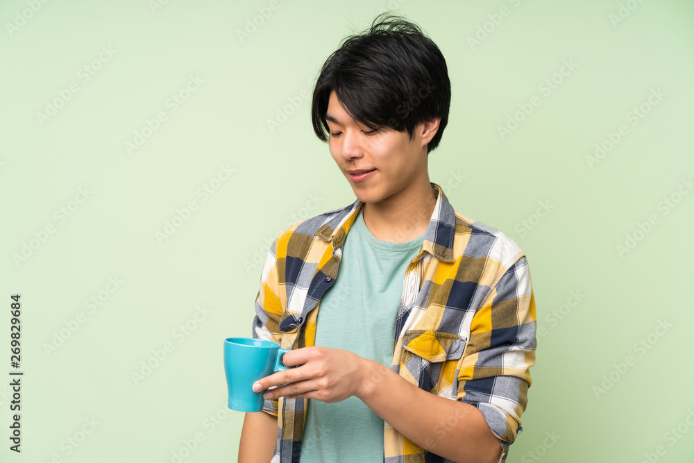 Asian man over isolated green wall holding hot cup of coffee Asian man over isolated green wall holding hot cup of coffee
