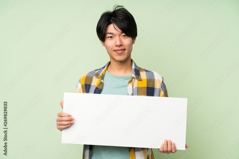 Asian man over isolated green wall holding an empty white placard for insert a concept Asian man over isolated green wall holding an empty white placard for insert a concept