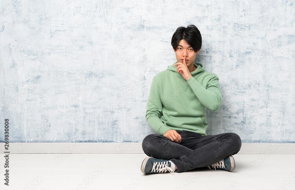Asian man sitting on the floor showing a sign of silence gesture putting finger in mouth